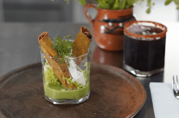 two taquitos placed in a glass cup serving dish with green salsa